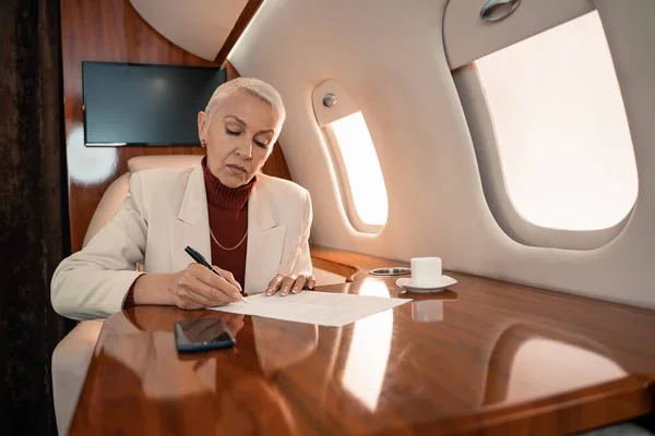 Зрелая деловая женщина, пишущая на бумаге возле размытого смартфона и кофе в самолете — стоковое фото