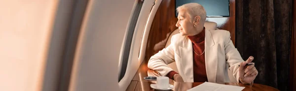 Усміхнена бізнес-леді дивиться на вікно літака біля кави і документа, банер — стокове фото