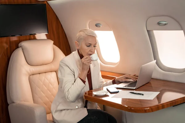 Businesswoman drinking coffee and using laptop near document in plane — Stock Photo