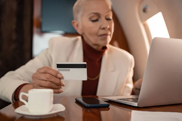 Credit card in hand of blurred businesswoman using laptop near coffee in plane — Stock Photo