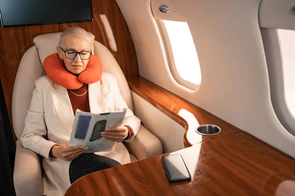 Mujer de negocios en la revista de lectura de almohadas cuello cerca de teléfono inteligente borroso en avión - foto de stock
