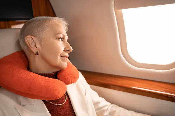Зрелая деловая женщина в подушке на шее смотрит в окно самолета — стоковое фото