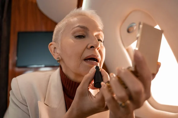 Mujer madura aplicando lápiz labial y sosteniendo espejo en chorro - foto de stock