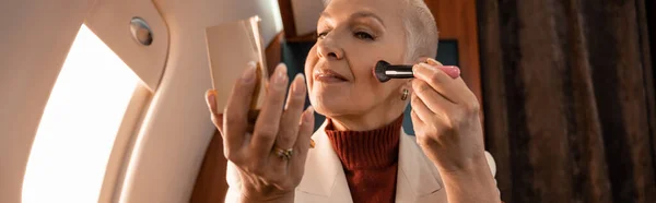 Mature woman applying blushes and holding mirror in plane, banner — Stock Photo