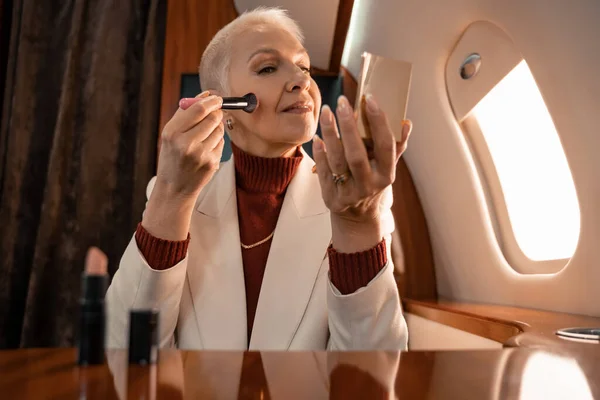 Mature woman applying blushes and holding mirror in airplane — Stock Photo