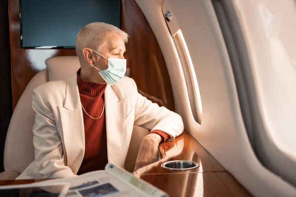 Mujer de negocios en máscara médica mirando la ventana del avión cerca de la revista y la tableta digital - foto de stock