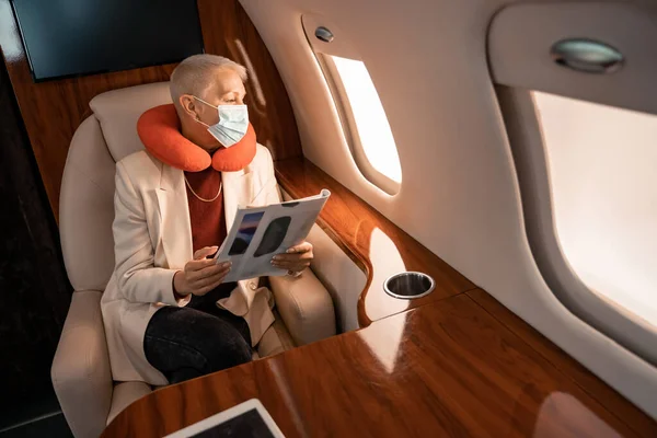 Geschäftsfrau mit Nackenkissen und medizinischer Maske hält Magazin in der Hand und blickt auf Flugzeugfenster — Stockfoto