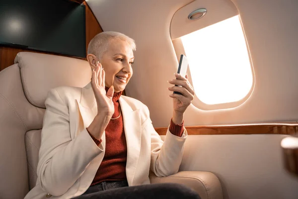 Улыбающаяся деловая женщина машет рукой во время видеозвонка на смартфон в самолете — стоковое фото