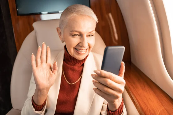 Femme d'affaires joyeuse ayant chat vidéo sur smartphone flou dans un avion privé — Photo de stock