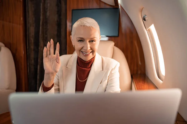 Mujer de negocios sonriente teniendo videollamada en un ordenador portátil borroso en avión - foto de stock