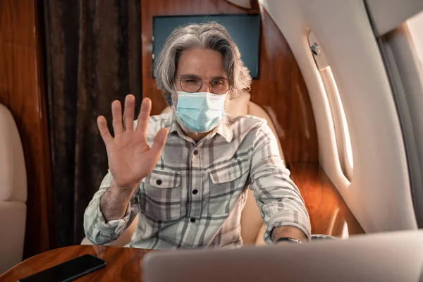 Empresário em máscara médica tendo videochamada no laptop em jato — Fotografia de Stock