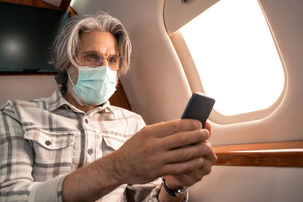 Man in medical mask using blurred smartphone in cabin of plane — Stock Photo