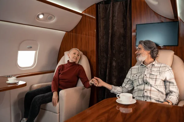 Smiling mature couple holding hands near cups of coffee in private plane — Stock Photo