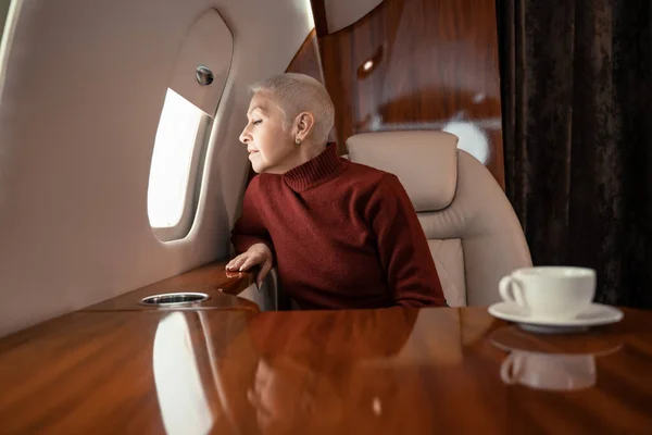 Зрелая женщина смотрит в окно частного самолета возле размытой чашки кофе — стоковое фото