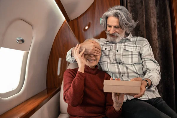 Sonriente hombre sosteniendo caja de regalo y cubriendo los ojos de la esposa en avión privado - foto de stock