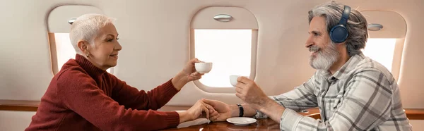 Vista lateral de la mujer sonriente sosteniendo la taza cerca del marido en los auriculares en el avión, bandera - foto de stock