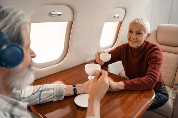 Улыбающаяся женщина держит чашку кофе рядом с размытым мужем в наушниках в частном самолете — стоковое фото