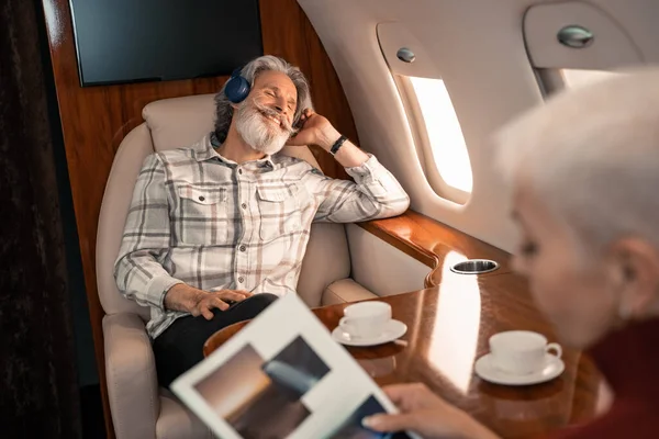 Sonriente hombre en los auriculares sentado cerca de la esposa leyendo revista en primer plano borrosa - foto de stock