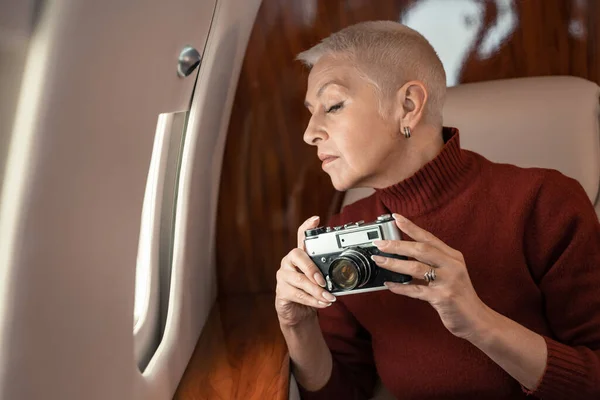 Mulher madura segurando câmera retro perto da janela no avião — Fotografia de Stock