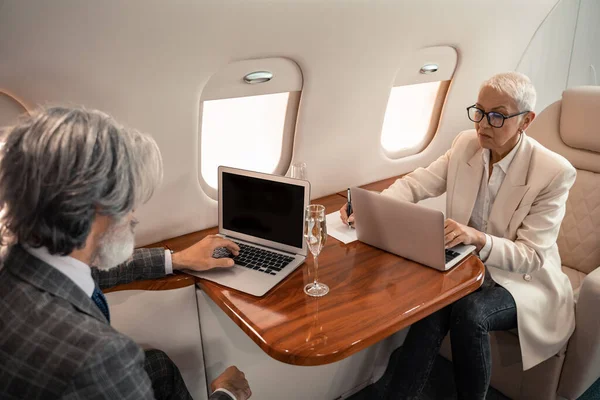 Donna d'affari matura che scrive su notebook e utilizza laptop vicino champagne e uomo d'affari in aereo — Foto stock