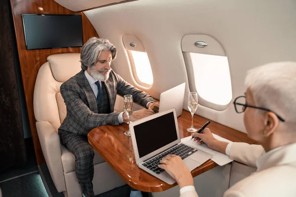 Geschäftsmann benutzt Laptop in der Nähe unscharfe Geschäftsfrau Schreiben auf Papier und Champagner im Privatjet — Stockfoto