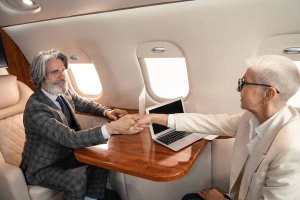 Улыбающийся бизнесмен пожимает руку бизнесвумен возле ноутбуков в самолете — стоковое фото