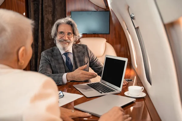 Улыбающийся бизнесмен, указывающий на ноутбук возле кофе и размытый бизнесмен в частном самолете — стоковое фото