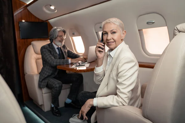 Empresária sorridente falando no smartphone e olhando para a câmera perto de empresário borrado no avião — Fotografia de Stock