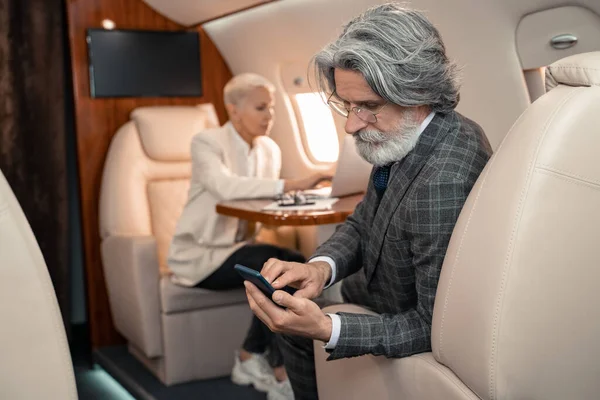 Бизнесмен, использующий смартфон рядом с размытой бизнес-леди в самолете — стоковое фото