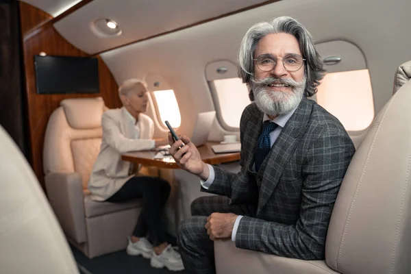 Lächelnder Geschäftsmann hält Smartphone in der Nähe von verschwommenem Geschäftspartner im Privatflugzeug — Stockfoto