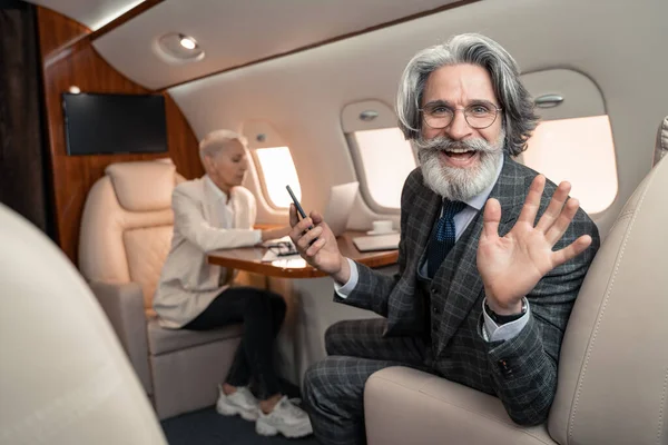 Uomo d'affari sorridente con smartphone che saluta la macchina fotografica vicino a donna d'affari offuscata in aereo — Foto stock