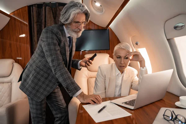 Businessman with smartphone looking at paper near businesswoman with laptop in private plane — Stock Photo