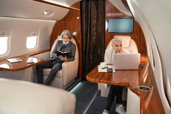 Businesswoman using laptop near coffee and partner with notebook in private plane — Stock Photo