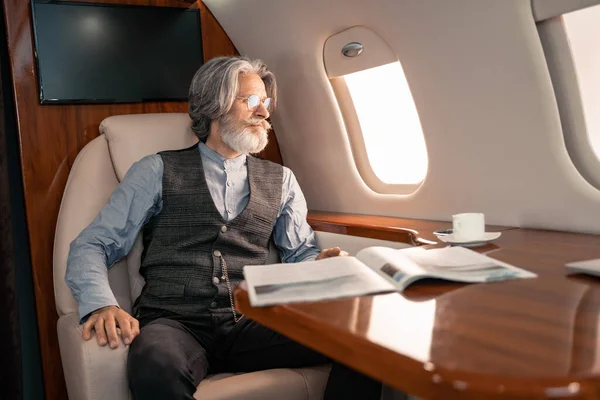 Uomo maturo guardando finestra vicino rivista e caffè in aereo — Foto stock