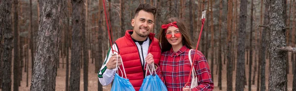 Feliz pareja con bolsas de basura sosteniendo recoger herramientas en el bosque, pancarta - foto de stock