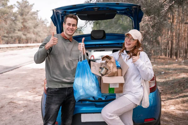 Voluntarios felices mostrando pulgares hacia arriba cerca del coche con basura - foto de stock