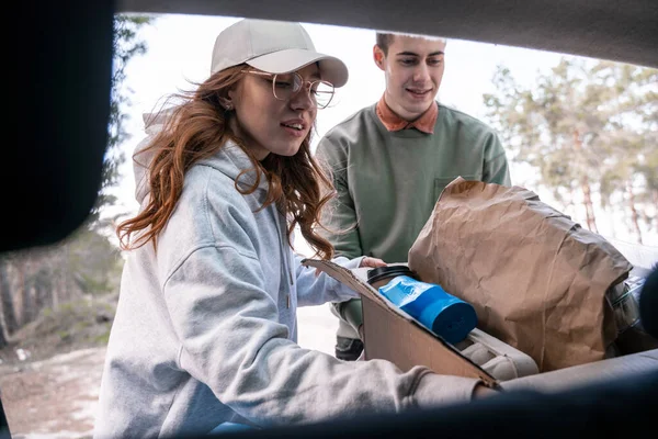 Молоді добровольці тримають картонну коробку зі сміттям біля машини — стокове фото
