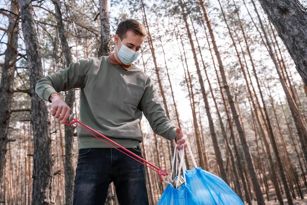 Voluntario en máscara médica sosteniendo recoger herramienta cerca de la basura en el bosque - foto de stock
