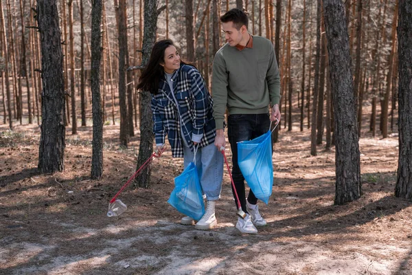 Feliz joven y hombre con bolsas de basura recogiendo basura con herramientas de agarre en el bosque - foto de stock