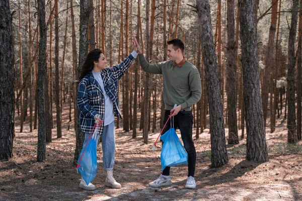 Comprimento total de casal feliz com sacos de lixo dando alta cinco enquanto pegando lixo com ferramentas grabber na floresta — Fotografia de Stock