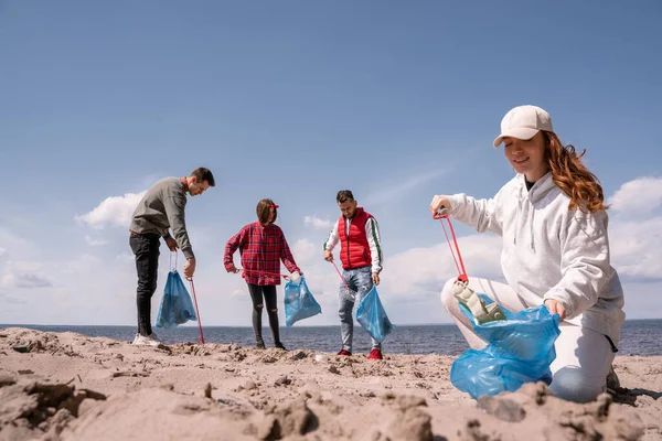 Glückliche Frau hält Müllsack in der Hand und sammelt Müll im Sand in der Nähe von Freiwilligen auf — Stockfoto