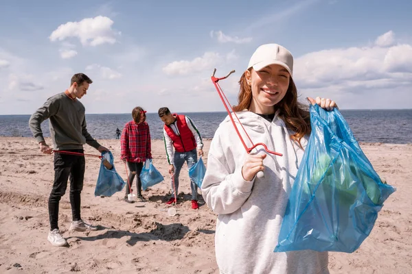 Mujer feliz sosteniendo la bolsa de basura y recoger herramienta cerca del grupo de voluntarios - foto de stock