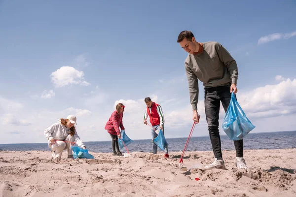 Hombre feliz sosteniendo la bolsa de basura y recogiendo basura en la arena cerca del grupo de voluntarios - foto de stock