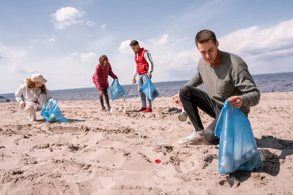 Joven sosteniendo la bolsa de basura y recogiendo basura en la arena cerca del grupo de voluntarios - foto de stock
