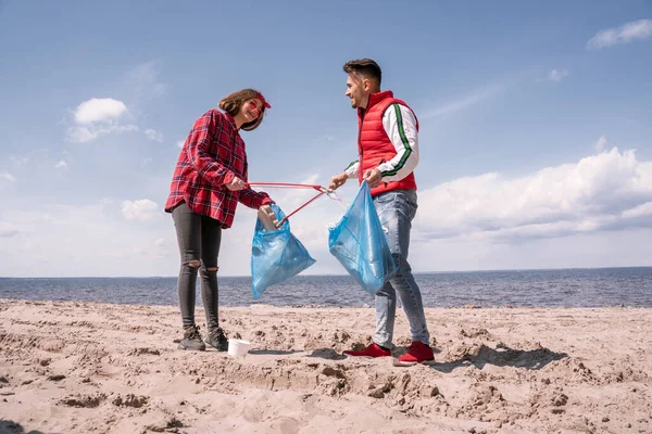 Happy couple with trash bags and grabbers picking up garbage on sand — Stock Photo