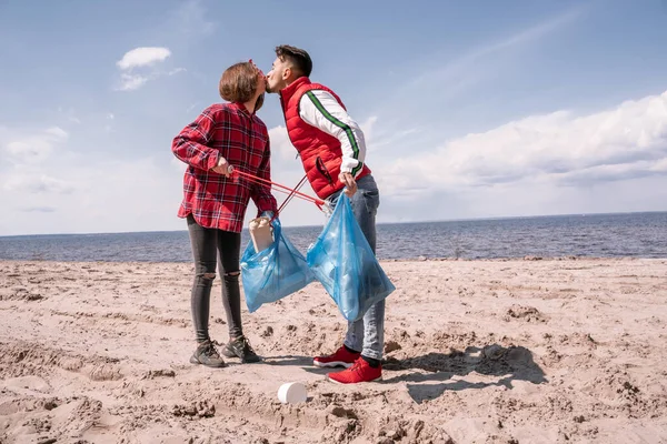 Paar mit Müllsäcken und Greifern küsst sich, während es auf Sand steht — Stockfoto