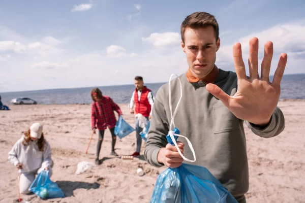 Joven mostrando gesto de stop cerca de grupo borroso de voluntarios recogiendo basura, concepto de ecología - foto de stock