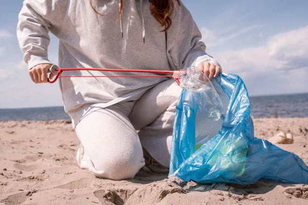 Vista recortada de la mujer sosteniendo bolsa de basura y recogiendo basura en la arena - foto de stock