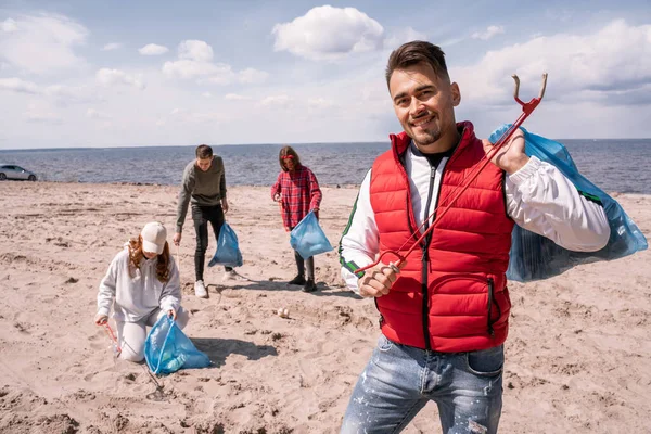 Fröhlicher Mann mit Müllsack und Greifer in der Nähe von verschwommenen Freiwilligen, die Müll auf Sand sammeln — Stockfoto