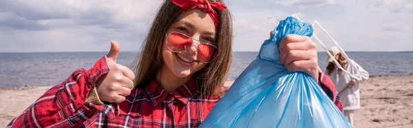 Mujer joven feliz en gafas de sol sosteniendo la bolsa de basura y mostrando el pulgar hacia arriba, pancarta - foto de stock